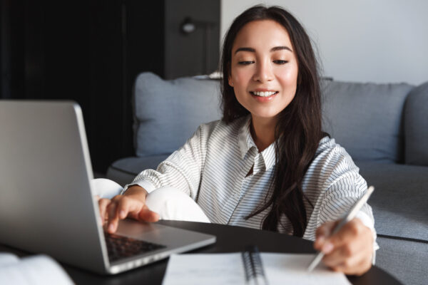 Image of beautiful asian girl studying remote from home, writing down lecture while using virtual classroom on laptop. Woman preparing for remote meeting, using computer.